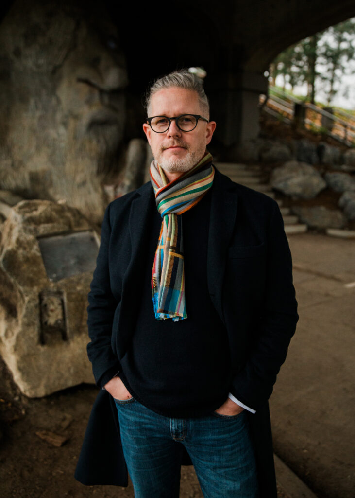 A portrait of Bill Hulseman, a middle-aged man with short gray hair, a neatly trimmed beard, and black-framed glasses. He is wearing a black coat over a dark sweater, complemented by a colorful striped scarf. His hands are casually tucked into his jean pockets as he stands confidently in front of a large stone sculpture of a troll under a bridge. The outdoor setting features earthy tones, stone steps, and trees in the background, creating a moody yet artistic atmosphere. His expression is calm and introspective, reflecting a sense of depth and creativity.