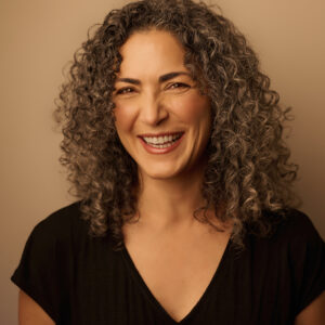 Atousa Raissyan, a life alchemist, smiles warmly in a professional portrait. She has curly, salt-and-pepper hair and wears a black V-neck top. Her radiant expression and slightly squinting eyes convey joy and authenticity. The background is a soft, warm beige, complementing her natural features. The lighting is soft and even, highlighting her face with a flattering glow.