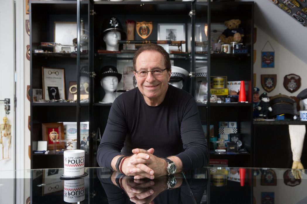 Bestselling author Peter James sits at a reflective glass table, smiling at the camera. He wears a black long-sleeve shirt, glasses, and a wristwatch. Behind him is a large shelving unit filled with police memorabilia, including helmets, badges, framed photos, plaques, and other law enforcement artifacts. A white coffee mug with "City of London Police" text is placed on the table in front of him. The room has a well-lit, organized display of law enforcement collectibles.